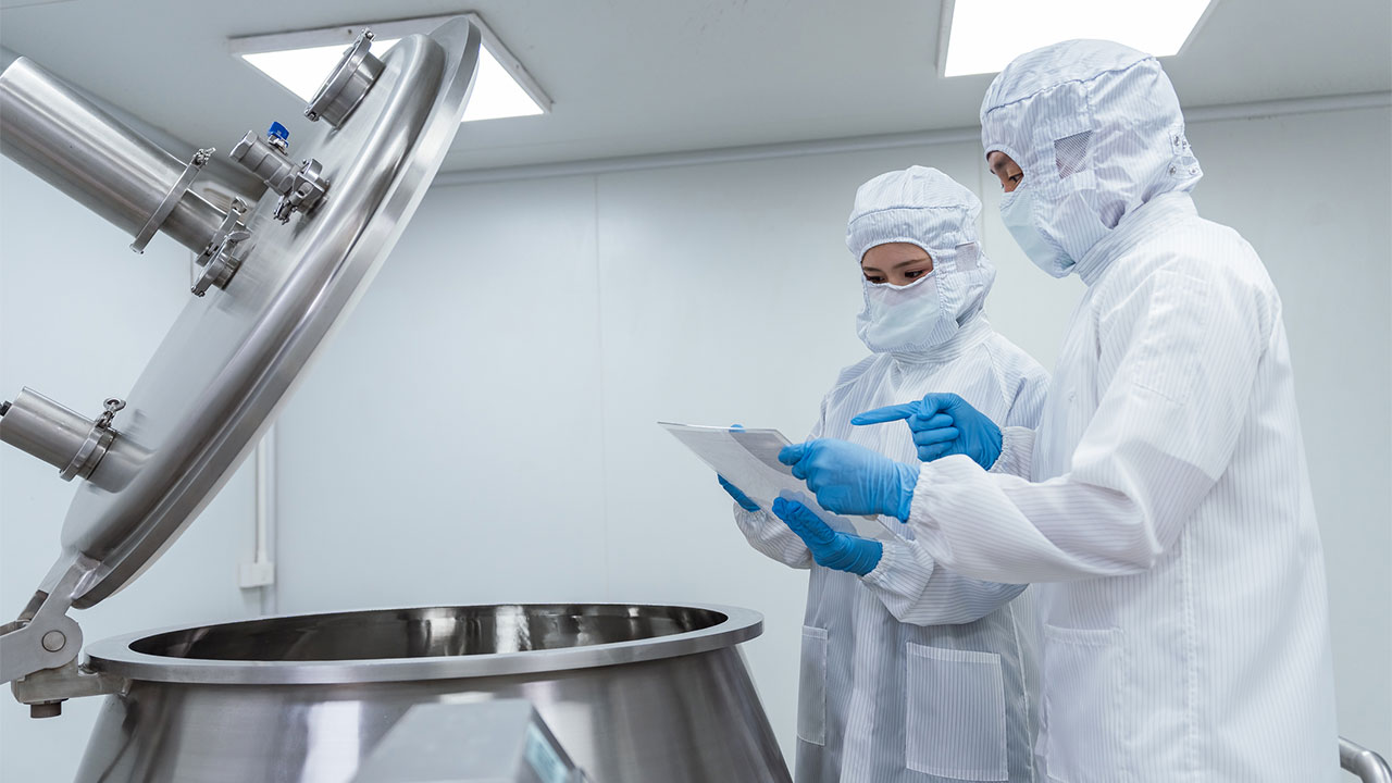 Two technicians wearing white protective suits and face masks inspecting a large stainless steel manufacturing still
