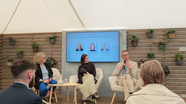 Three panel members sitting on a raised stage with a presentation screen behind and audience members in the foreground